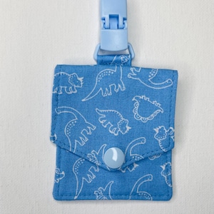 May include: A light blue pacifier clip with a square pouch featuring a white dinosaur pattern. The pouch has a light blue button closure and a matching clip.