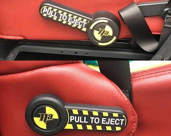 May include: Black and yellow "PULL TO EJECT" lever with a logo, set against a red seat. The lever is attached to a black base. The text is in white on a black and yellow striped background.