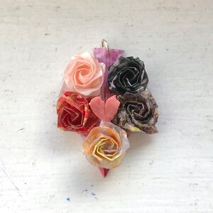 以下が含まれることがあります： ピンク、赤、黄色、黒、金色の折り紙の花が、紫色の背景にハート型に配置されたペンダントです。