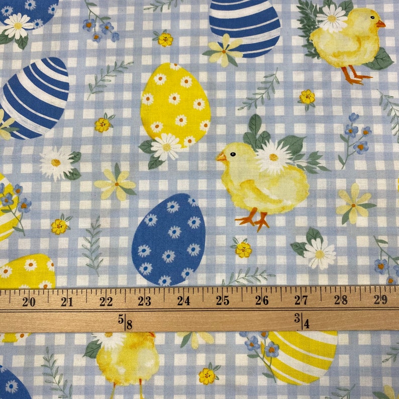 Puede incluir: Huevos de Pascua azules y amarillos y pollitos amarillos sobre un fondo de cuadros azules y blancos. Una regla de madera con marcas en pulgadas est&aacute; en la parte inferior de la imagen.