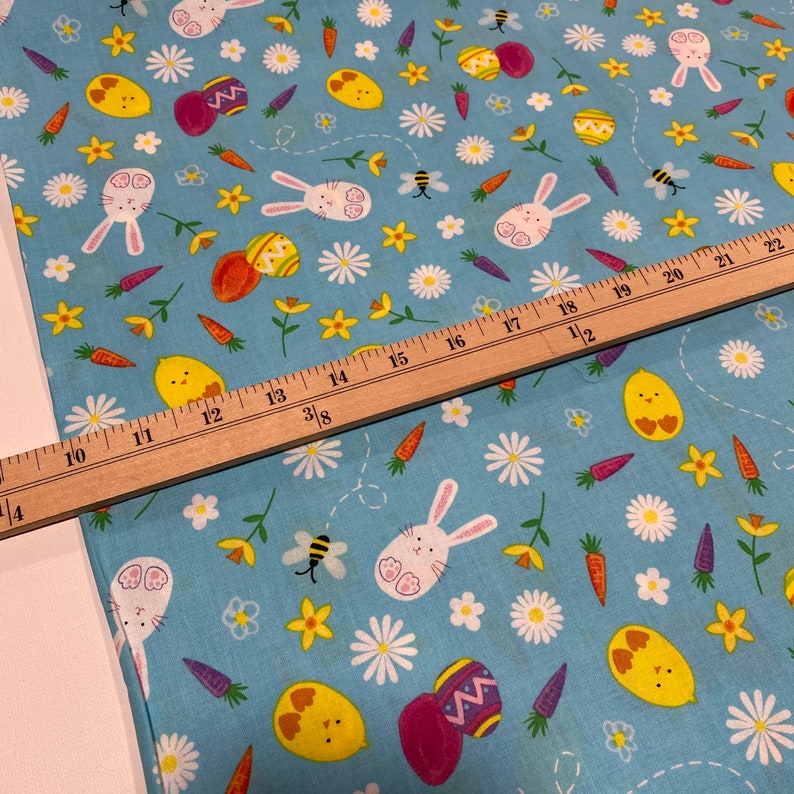 Puede incluir: Una tela azul con un patr&oacute;n repetido de im&aacute;genes con tem&aacute;tica de Pascua, incluyendo conejos, pollitos, zanahorias, huevos y flores. La tela se mide con una regla.