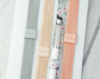 May include: Four elastic hair ties in gray, peach, floral print, and peach. A white pen with a clear crystal design is placed in the center of the hair ties.