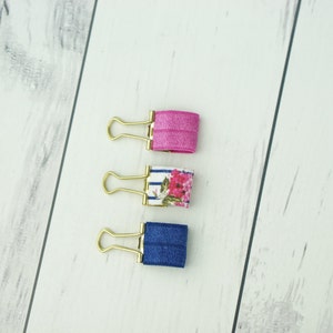May include: Three gold binder clips with fabric covers. The top clip is pink, the middle clip has a white and blue striped pattern with pink flowers, and the bottom clip is blue.