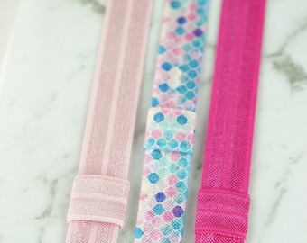 May include: Three hair ties, one pink, one pink with a blue and white geometric pattern, and one hot pink.