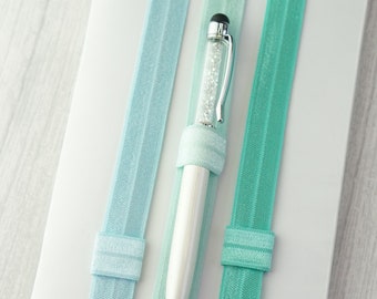 May include: A white notebook with three light blue elastic bands. The notebook has a silver pen with a clear body filled with glitter.
