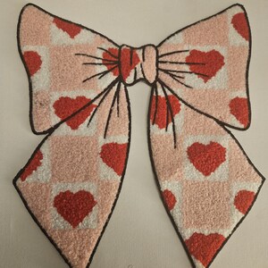 Può includere: Un grande fiocco decorativo con un motivo a quadri rosa e bianco, impreziosito da cuori rossi. Il fiocco ha un contorno nero ed è posto su uno sfondo bianco. Il design del fiocco ricorda San Valentino.