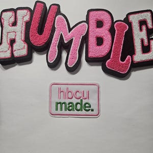 May include: A chenille patch spelling the word HUMBLE in alternating white and pink letters with a black border, positioned above a rectangular white embroidered patch featuring the pink text hbcu and green text made. with a pink border.