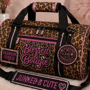 Op de afbeelding: Een duffeltas met luipaardprint, zwarte banden en roze strass-accenten. De tas is voorzien van geborduurde teksten met Blessed & Bougie, Fashion Junkie, Expensive and Difficult en Junked & Cute op de schouderband.