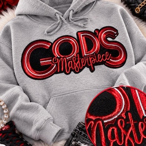 May include: A heather gray hooded sweatshirt featuring a large, embroidered patch on the chest with the text "GOD'S Masterpiece" in bold, metallic red lettering outlined in white against a black background, including a circular detail view.