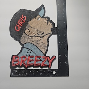 Könnte beinhalten: Ein gestickter Aufnäher mit einem Profilbild einer Person, die eine schwarze Mütze mit dem roten Schriftzug CHRIS und ein blaues Hemd trägt, darunter das Wort BREEZY in Rot. Ein schwarzes L-förmiges Lineal zeigt, dass der Aufnäher 20 cm hoch ist.
