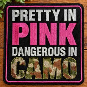 May include: A square embroidered patch with a black background and a pink border. The text reads PRETTY IN in silver, PINK in bright pink, DANGEROUS IN in silver, and CAMO in a green, brown, and tan camouflage pattern with a black outline.