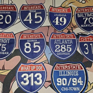 May include: A collection of ten embroidered shield-shaped patches featuring red, white, and blue designs. The patches display various text including Interstate, state names like Louisiana, Texas, Georgia, Michigan, Illinois, and city names like Baltimore, Detroit, Atlanta, and Chi-Town, along with route numbers.