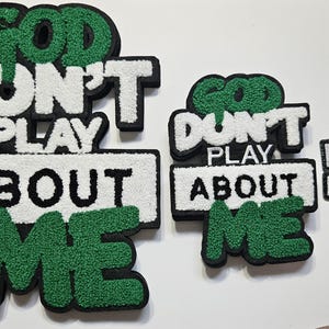 May include: Three tufted wall art pieces in descending sizes, each featuring the text GOD DON'T PLAY ABOUT ME. The letters are rendered in a combination of green, white, and black yarn with a textured, raised surface and a thick black border.