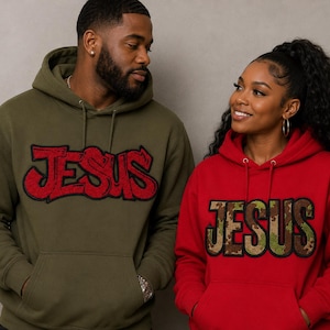 Puede incluir: Una sudadera con capucha verde oliva con un parche rojo de JESUS estilo grafiti y una sudadera roja con un parche de JESUS con patrón de camuflaje. Ambas prendas tienen mangas largas, un bolsillo tipo canguro y capucha con cordón.