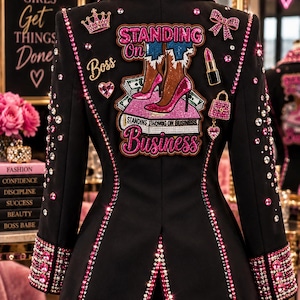 May include: A black blazer embellished with pink and clear rhinestones along the collar, cuffs, and seams. The back features a large embroidered patch with the text "STANDING ON BUSINESS" and an illustration of legs in pink heels on money.
