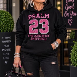 Könnte beinhalten: Ein schwarzer Kapuzenpullover mit dem rosa Text PSALM 23 und THE LORD IS MY SHEPHERD sowie einer großen, glitzernden rosa Zahl 23. Der Hoodie verfügt über eine Kängurutasche vorne und lange Ärmel. Eine schwarze Handtasche mit dem Text Grandma with Grace & Grit ist ebenfalls abgebildet.