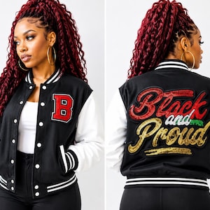May include: A black and white varsity jacket featuring a red letter B patch on the front left chest. The back displays large, decorative sequined text that reads Black and Proud in red, green, and gold colors, with striped ribbed trim on the cuffs.
