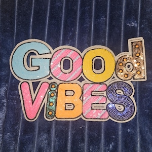 May include: A decorative patch featuring the text GOOD VIBES in large, multi-colored letters. The letters are embellished with sequins and beads in shades of light blue, pink, yellow, gold, orange, and dark blue, outlined with a white border.