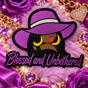 May include: An embroidered patch featuring a person wearing a purple hat with a white band, large yellow hoop earrings, and red lips. Below the figure, the text Blessed and Unbothered is stitched in purple cursive script on a black background.