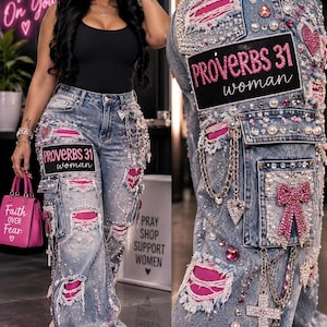 May include: Light blue denim cargo trousers featuring distressed sections with bright pink fabric underneath, embellished with pearls, rhinestones, pink gems, silver chains, cross charms, and a black patch with the text PROVERBS 31 woman.