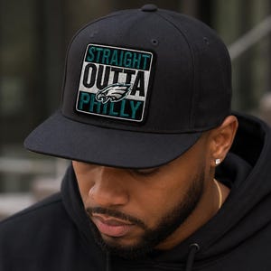 May include: A black baseball cap featuring a square patch on the front. The patch displays the text STRAIGHT OUTTA PHILLY in white and teal lettering, with a stylized eagle logo positioned between the words OUTTA and PHILLY.