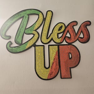 May include: A decorative sign with the words "Bless Up" in a colorful, sequined design. The word "Bless" is in green, yellow, and red, while "Up" is in yellow and red. The sign has a black outline.