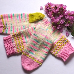 Könnte beinhalten: Ein Paar handgestrickte Socken aus einem bunten, gesprenkelten Garn in den Farben Rosa, Grün und Weiß. Die Socken haben einen rosa Bund und ein grün-weißes Muster.