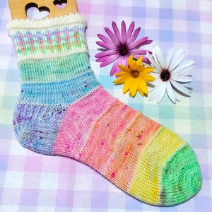 May include: A colorful hand-knitted sock with a green, yellow, orange, pink, blue, and white striped pattern. The sock has a white cuff with a small floral design.