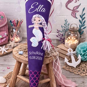 Könnte beinhalten: Eine lila-rosa Schultüte mit Meerjungfrauen-Motiv, dem Namen "Ella" und dem Text "Schulkind 15.08.2025". Die Tüte zeigt eine Meerjungfrau-Illustration und einen dekorativen Aufsatz mit dem Text "Du bist 1. Klasse".