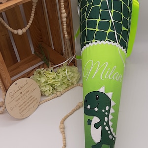 Puede incluir: Cono escolar verde y amarillo con un diseño de dinosaurio y el nombre "Milan" bordado en él. El cono tiene un borde blanco festoneado y el texto "Schulkind 2024" está impreso en la parte inferior.