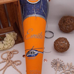 Può includere: Un cono scolastico arancione e blu con il nome "Emilio" in argento. Sotto il nome c'è una grafica di un elicottero della polizia. Il cono ha una parte superiore a fantasia blu e arancione con il testo "Du bist 1. Klasse" su un emblema circolare.