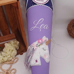 Peut inclure: Un cornet scolaire violet avec un motif de licorne et le nom "Lea" en écriture blanche. Le cône a un dessus bleu foncé avec une étiquette qui dit "Du bist 1. Klasse". Un article décoratif pour la rentrée scolaire.