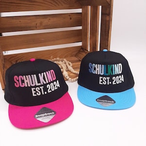 Op de afbeelding: Twee zwarte snapback caps met de tekst "SCHULKIND EST. 2024". Eén cap heeft een roze klep en de andere een blauwe klep. De caps worden gepresenteerd op een wit oppervlak.