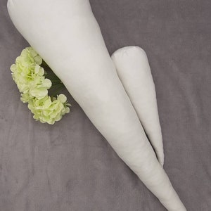 Könnte beinhalten: Zwei weiße, kegelförmige Kissen aus Stoff, eines größer als das andere, auf einer grauen, flauschigen Oberfläche. Ein kleiner Strauß hellgrüner Blumen befindet sich links neben den Kissen.
