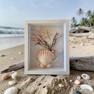 May include: A framed artwork composed of seashells, including a large, fan-shaped shell, a starfish, and smaller shells, arranged against a neutral backdrop. The white frame displays the beach scene in the background. The artwork is a decorative piece.