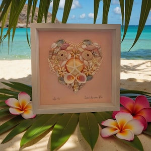 May include: A framed artwork with a heart design made from seashells, starfish, and sea urchins on a pink backdrop. The white framed artwork has text at the bottom. Tropical flowers and palm leaves are in the foreground, with a beach scene in the background.