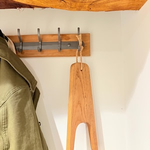 May include: A wooden boot jack with a natural wood finish hangs from a hook on a wooden wall. The boot jack is shaped like a wide A with a rounded top and a cutout in the middle.