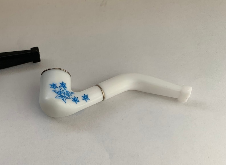 May include: White ceramic pipe with a blue floral design. The pipe has a curved stem and a round bowl.