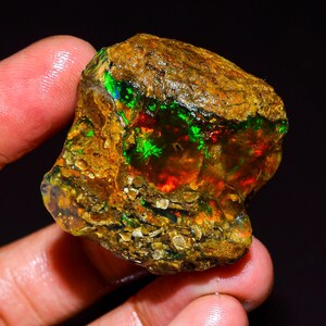 May include: A rough, uncut opal gemstone with a vibrant display of color. The stone features flashes of green, red, and blue within a brown matrix. The opal is held in a hand, showcasing its natural, raw form.