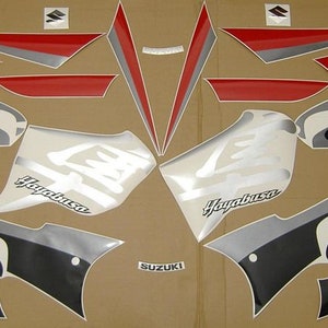 Puede incluir: Un conjunto de calcomanías para motocicleta en negro, rojo, plateado y blanco. Las calcomanías presentan las palabras "Hayabusa" y "GSX 1300" y el logotipo de Suzuki.