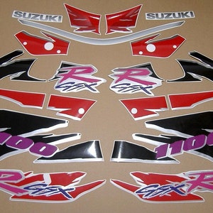 Puede incluir: Un conjunto de calcomanías para motocicleta en negro, rojo, plateado y morado. Las calcomanías presentan las palabras "Suzuki", "GSX-R" y "1100".
