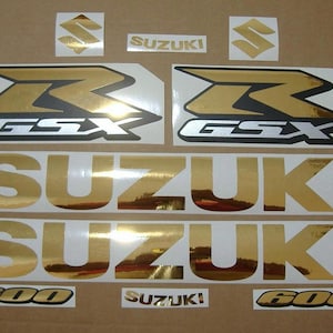 Puede incluir: Calcomanías de motocicleta doradas y plateadas con el texto "GSX" y "SUZUKI" en una fuente en negrita. Las calcomanías son para una motocicleta Suzuki GSX 600.