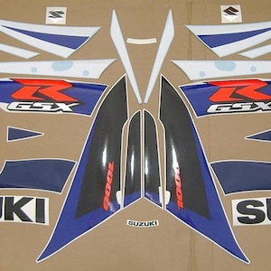 以下が含まれることがあります： スズキGSX 1000オートバイ用デカールのセット。デカールは主に青、白、黒、赤です。「SUZUKI」の文字が白いブロック体で表示されています。「GSX」のロゴも表示されています。