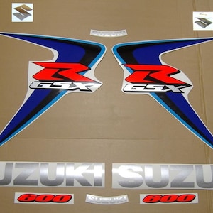 Puede incluir: Un juego de calcomanías para motos Suzuki GSX 600. Las calcomanías presentan una "R" roja y el texto "GSX", con detalles en azul y blanco. La palabra "SUZUKI" es plateada y "600" es roja.