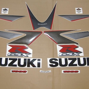 Puede incluir: Un conjunto de calcomanías para motocicleta en negro, rojo, plateado y blanco. Las calcomanías presentan las palabras "Suzuki" y "GSX 1000".