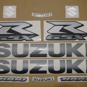 Può includere: Un set di adesivi per moto argentati e neri con il testo "GSX-R 1000" e "SUZUKI".
