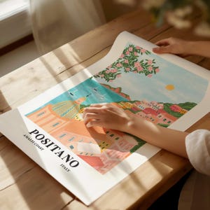 Puede incluir: Un p&oacute;ster ilustrado colorido con el texto "Positano, Amalfi Coast, Italy". El p&oacute;ster presenta una vista de la ciudad de Positano con edificios, una c&uacute;pula y un &aacute;rbol en flor.