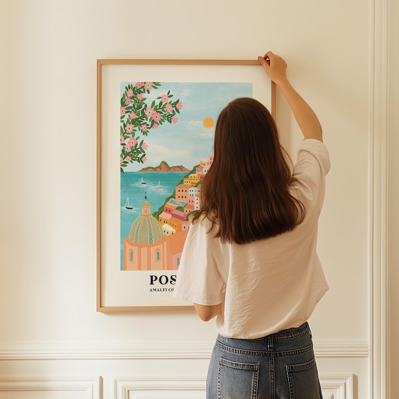 Puede incluir: Una ilustraci&oacute;n enmarcada de una ciudad colorida con una iglesia rosa, agua azul y un arbusto de flores rosas. El texto "Positano Amalfi Coast" est&aacute; en la parte inferior de la ilustraci&oacute;n.