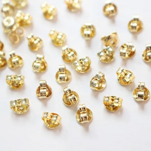 May include: A close-up of a group of gold-colored earring backs scattered on a white surface. The backs are designed to secure earrings to the earlobe.
