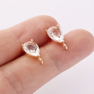 May include: Two gold-colored, pear-shaped jewelry findings with clear, faceted crystal stones. Each finding has a small loop at the bottom and a small prong at the top for attaching to other jewelry components. The findings are displayed against a neutral background.